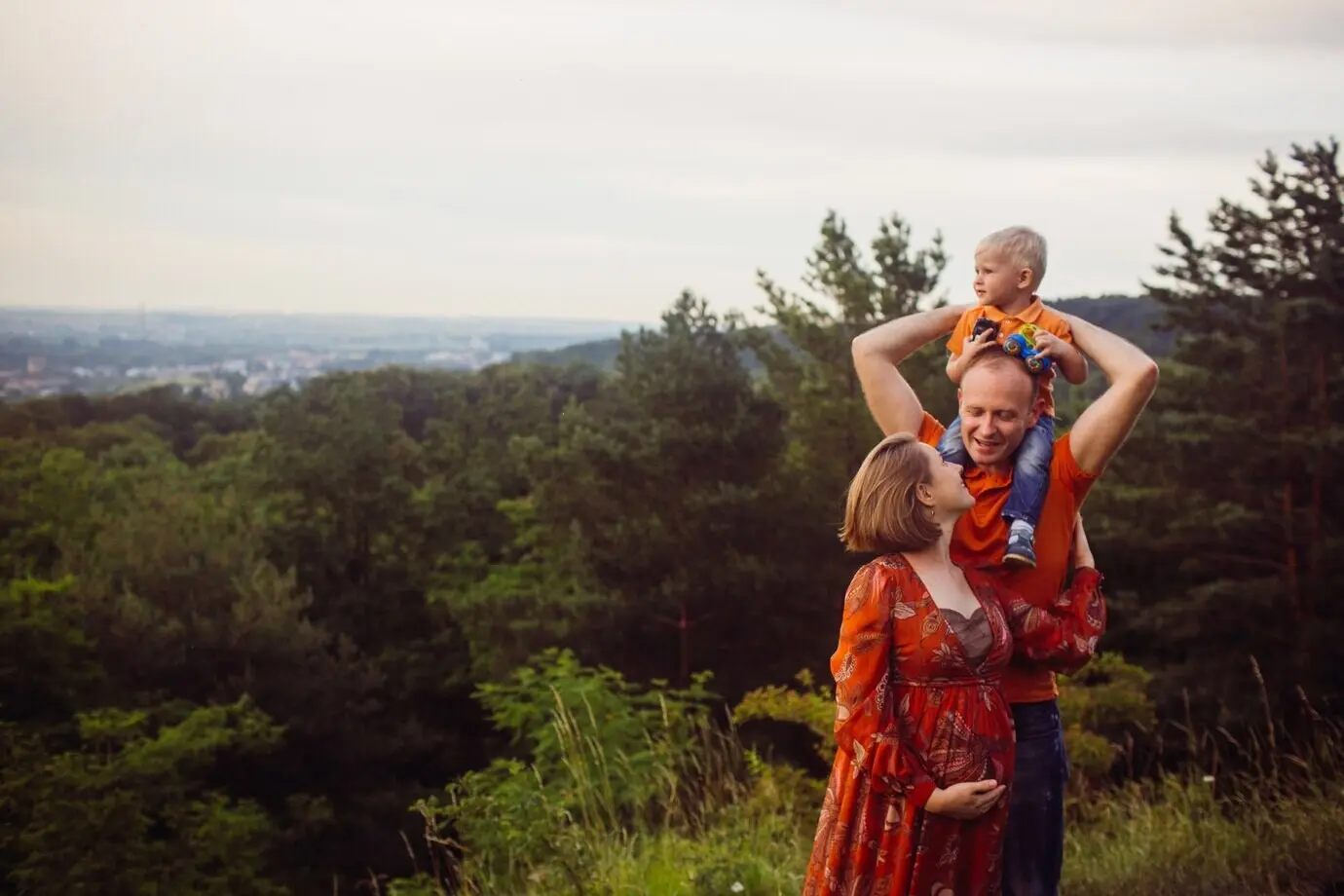 A father holds his son in his arms, posing with a pregnant woman in the forest.